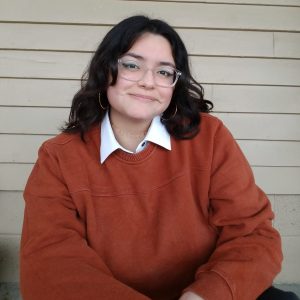 A photo of Arianna wearing a dark orange sweater and glasses.